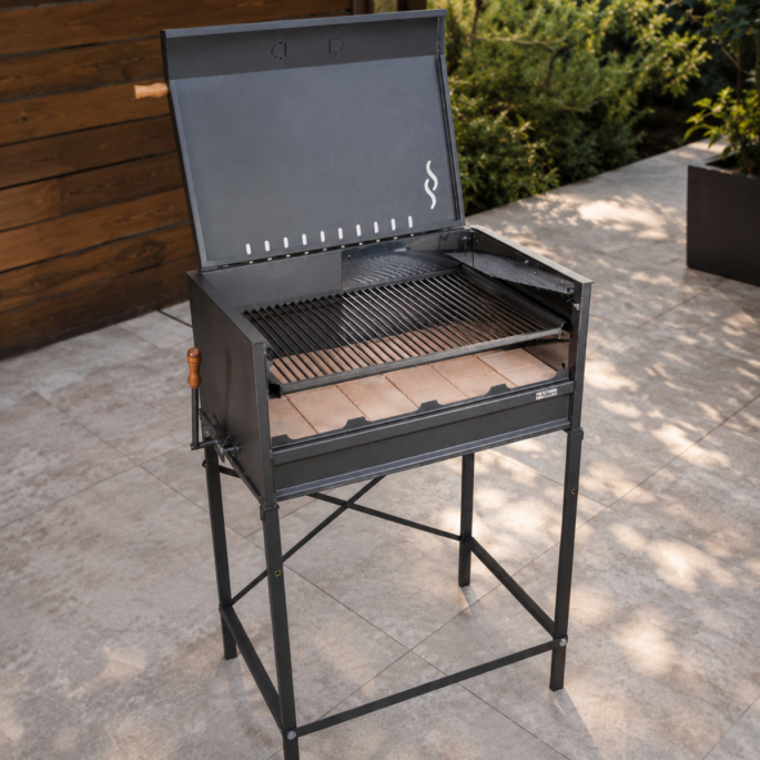 Nuke Pampa Argentijnse houtskoolbarbecue met open deksel, verstelbaar grillrooster en vuurvaste stenen op een betegeld terras.