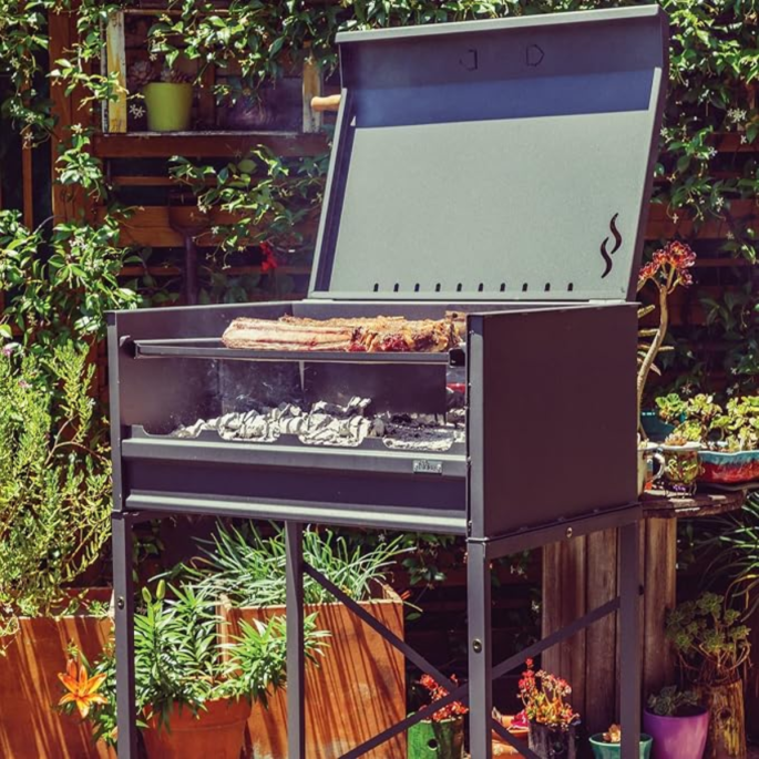 Nuke Pampa Argentijnse houtskoolbarbecue met open deksel, gegrild vlees en gloeiende houtskool in een groene tuin.